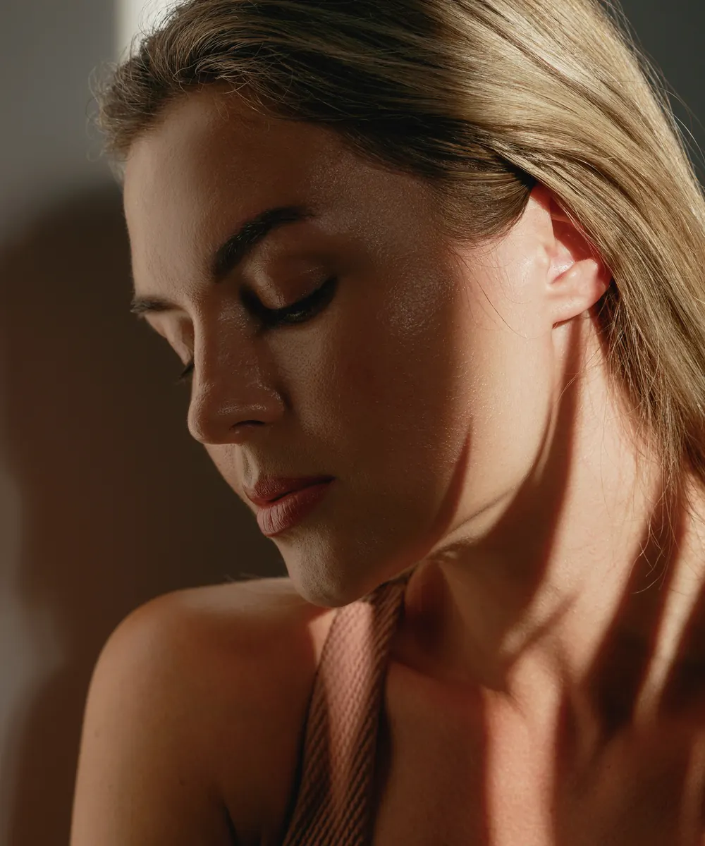 Blonde woman looks down as soft window light falls across her cheek and neck