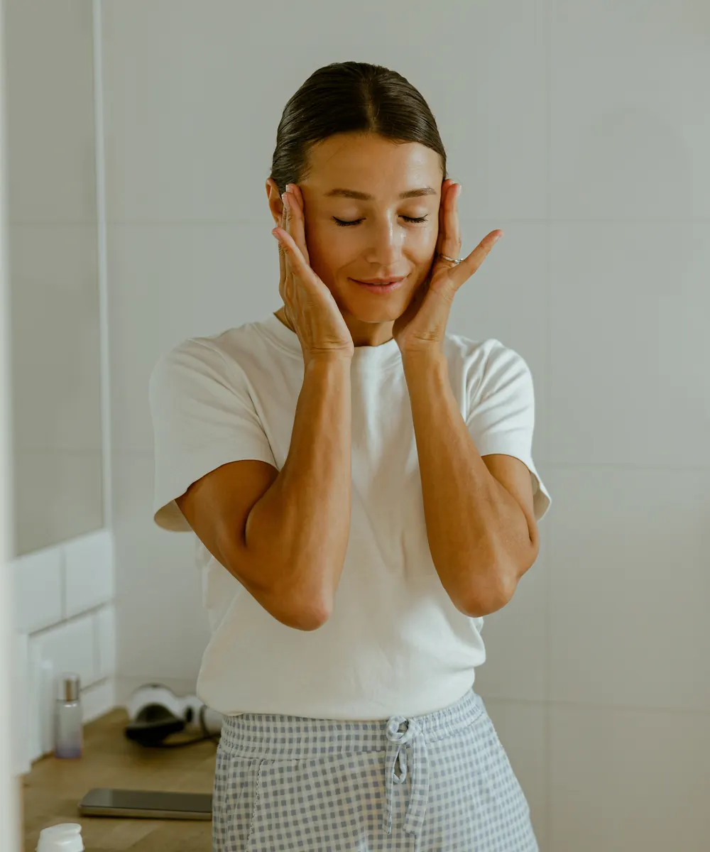 Woman in a white tee gently holds her face with both hands in a bright bathroom
