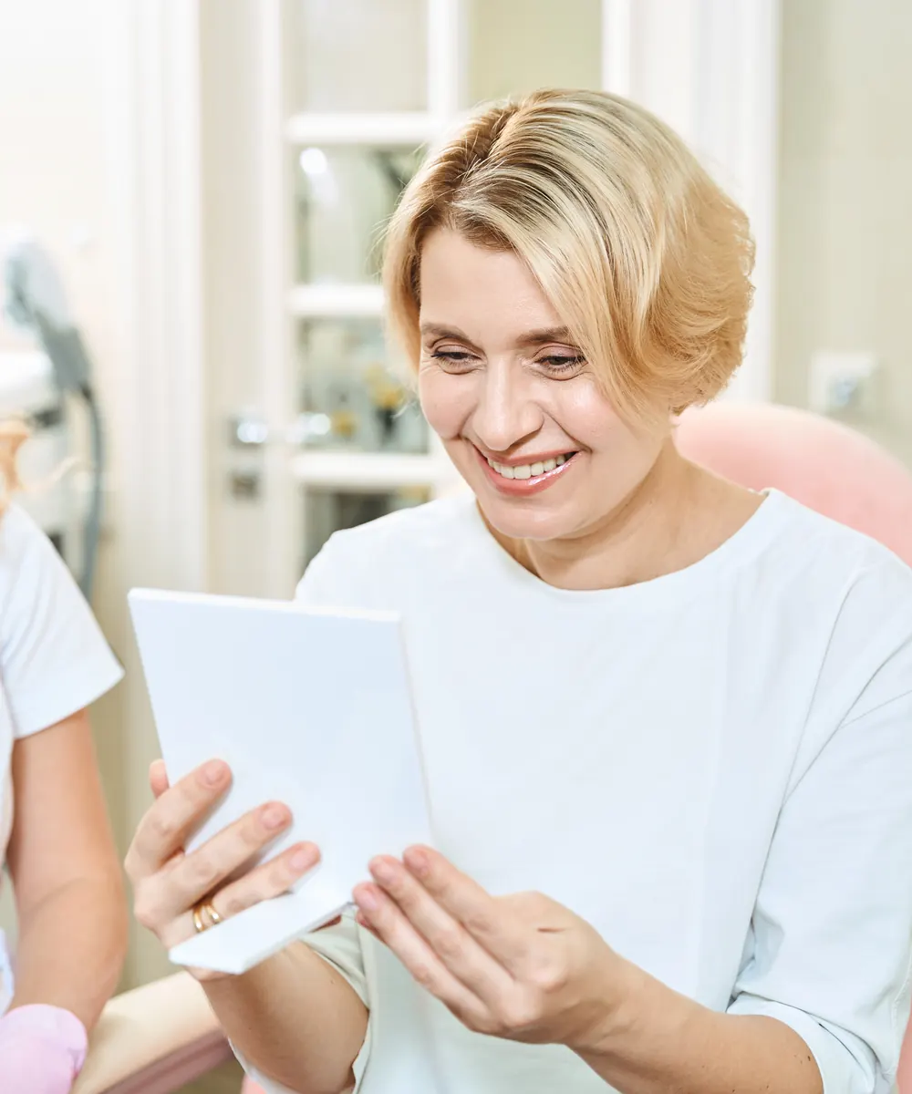 Smiling woman holds a hand mirror during a clinic visit with a practitioner beside her