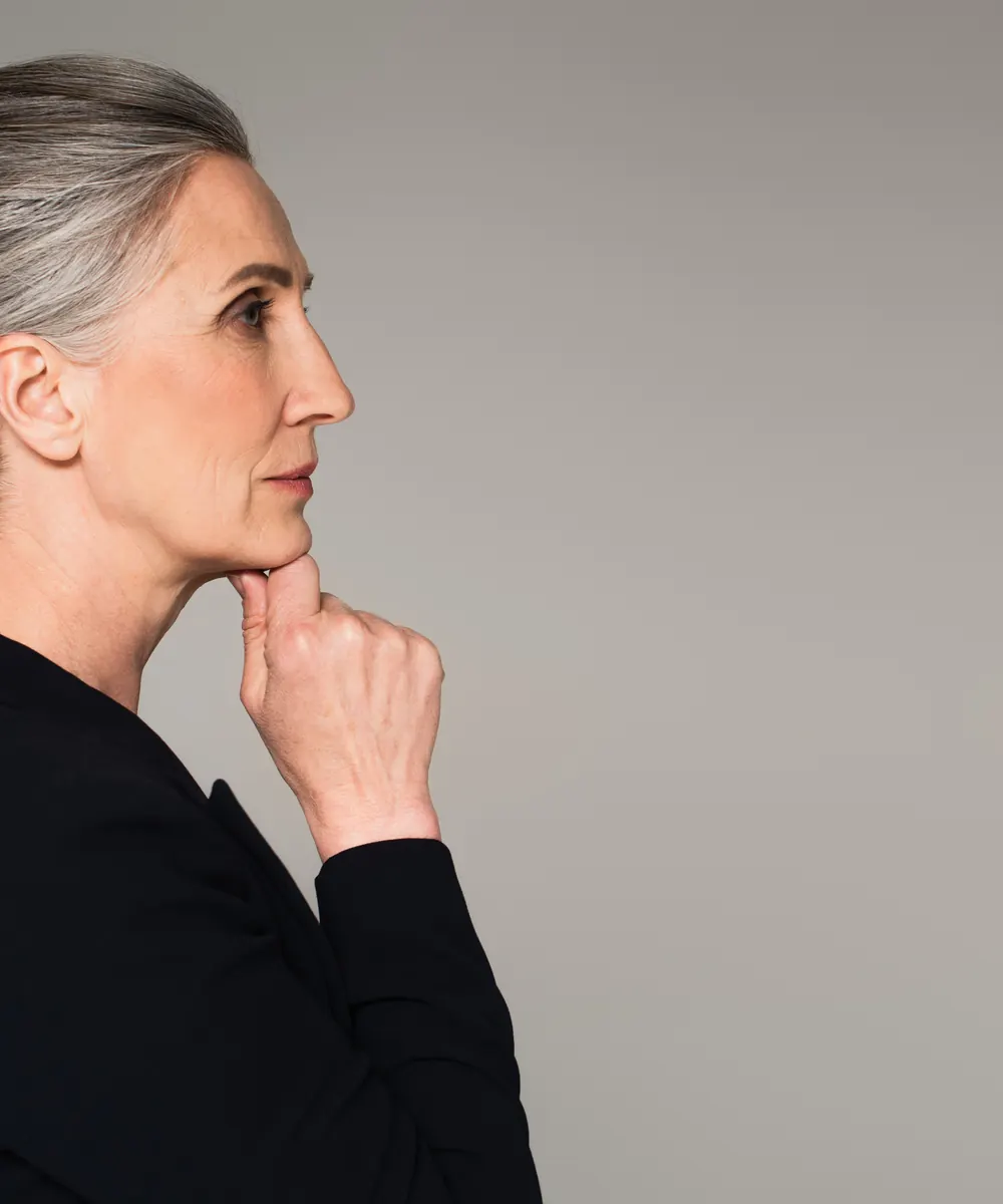 Grey haired woman in a black jacket stands in profile with her hand under her chin