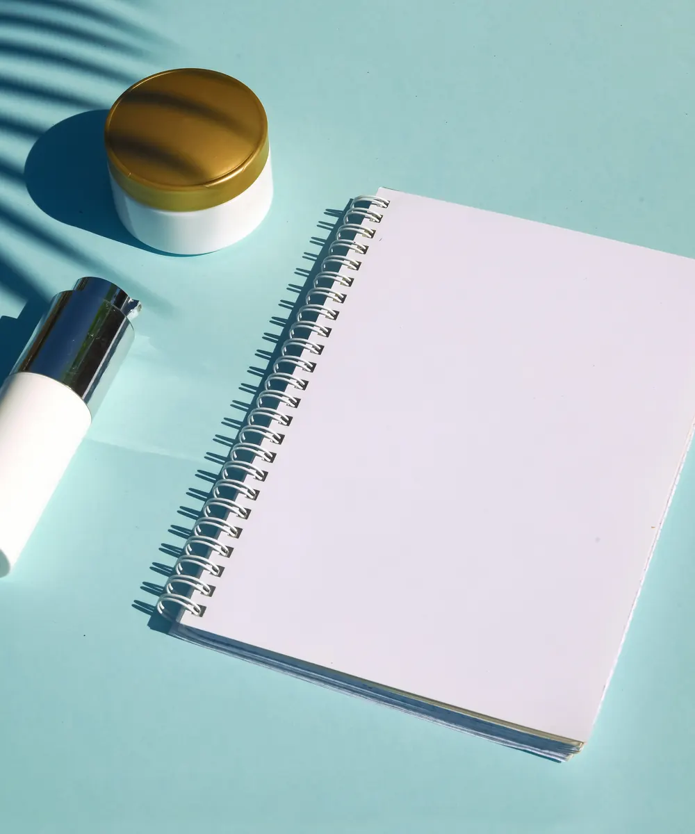 Blank spiral notebook beside a cream jar and serum bottle on a pale blue surface