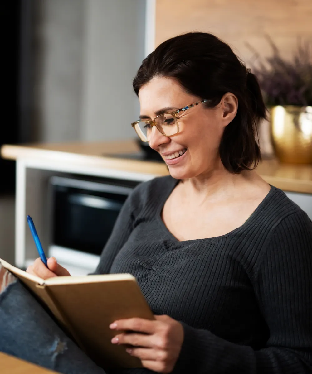 Smiling woman in glasses writes in a notebook while sitting in her kitchen