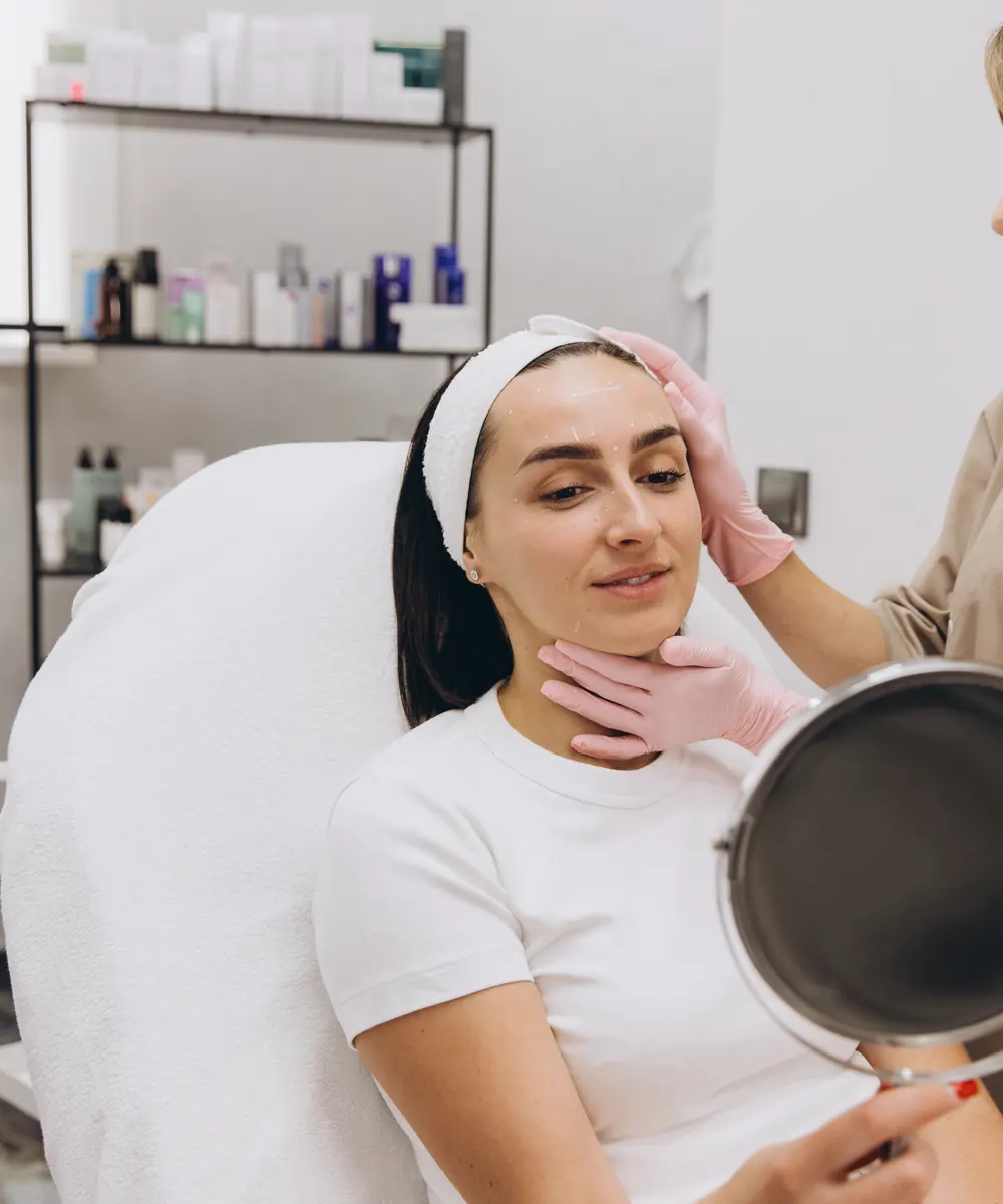 Woman in a treatment chair holds a hand mirror while a gloved clinician assesses her face