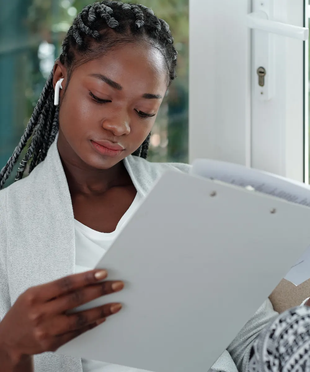 Woman with long braids reads a form on a clipboard while seated on a sofa