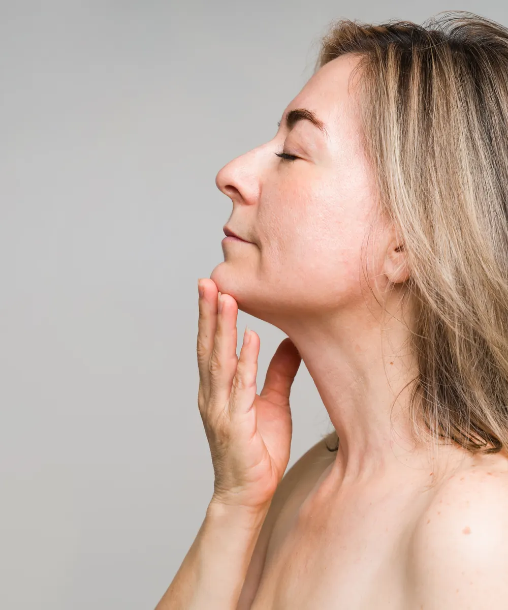 Woman in profile with eyes closed rests her hand along her jaw against a grey wall