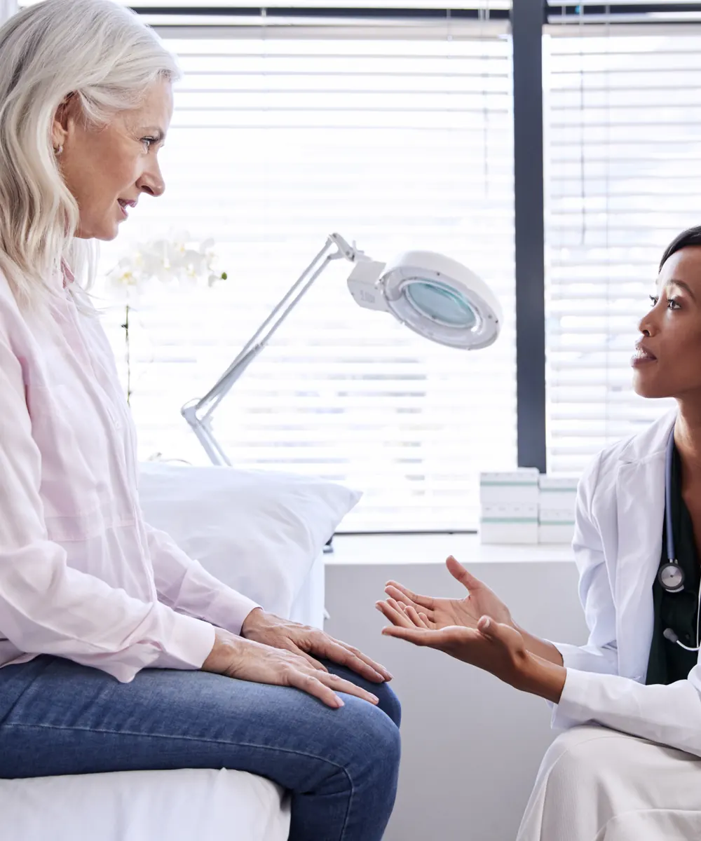 Woman with long grey hair sits on a treatment bed talking with a clinician