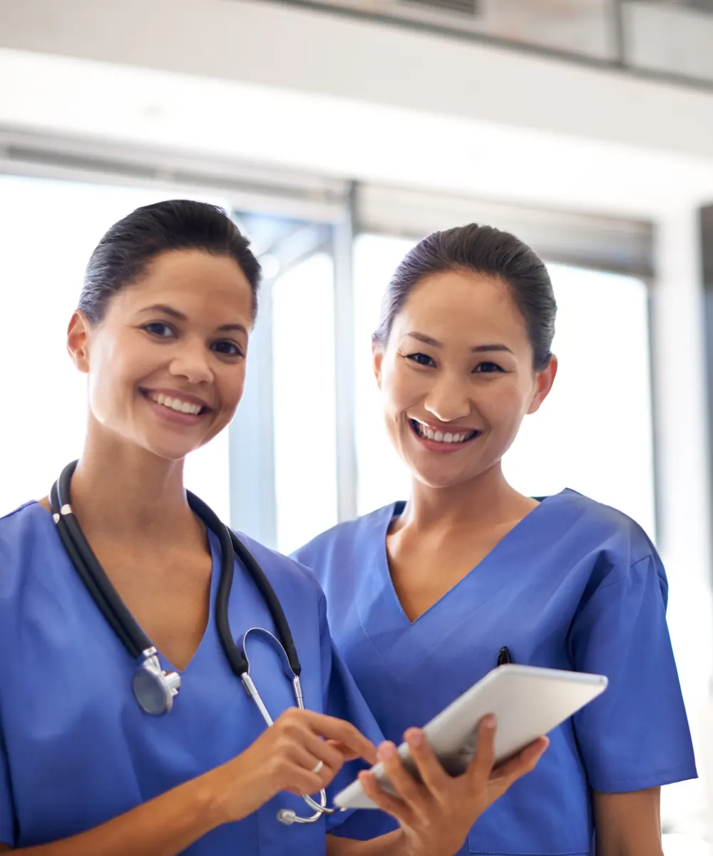 Two smiling nurses in blue scrubs, one wearing a stethoscope and holding a tablet