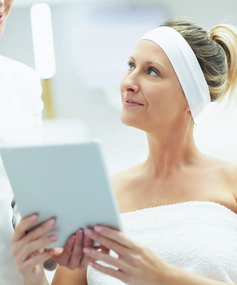 Woman in a white headband and towel looks up while holding a tablet in a clinic