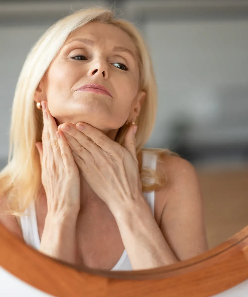 Mature blonde woman touches her jaw and neck while looking into a round mirror